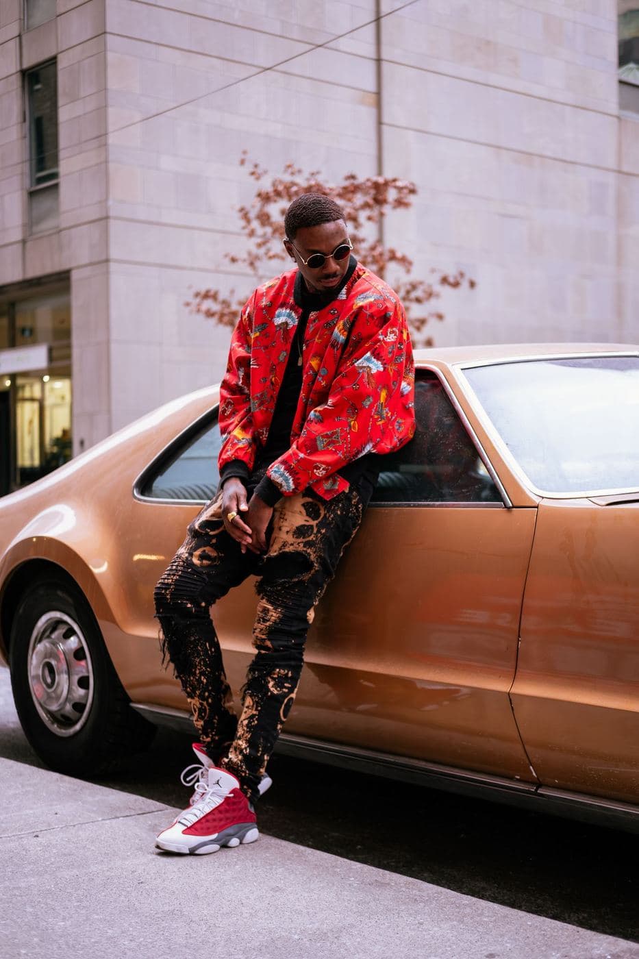 Man leaning on a bronze vintage car in a red patterned bomber jacket and matching trousers