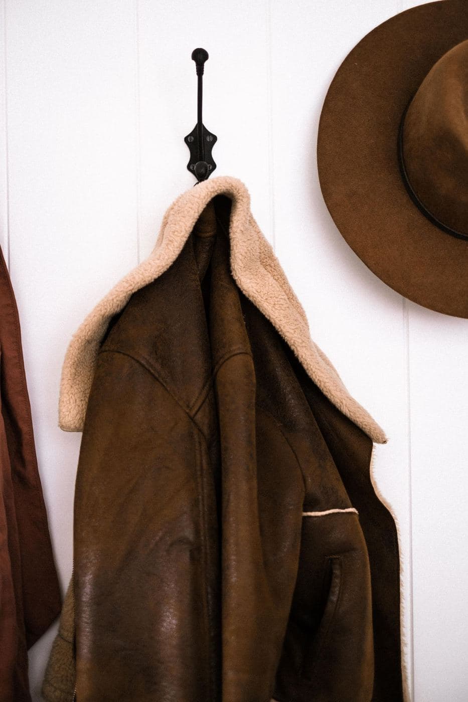 Brown suede sheepskin coat with a shearling collar hanging on a hook next to a hat