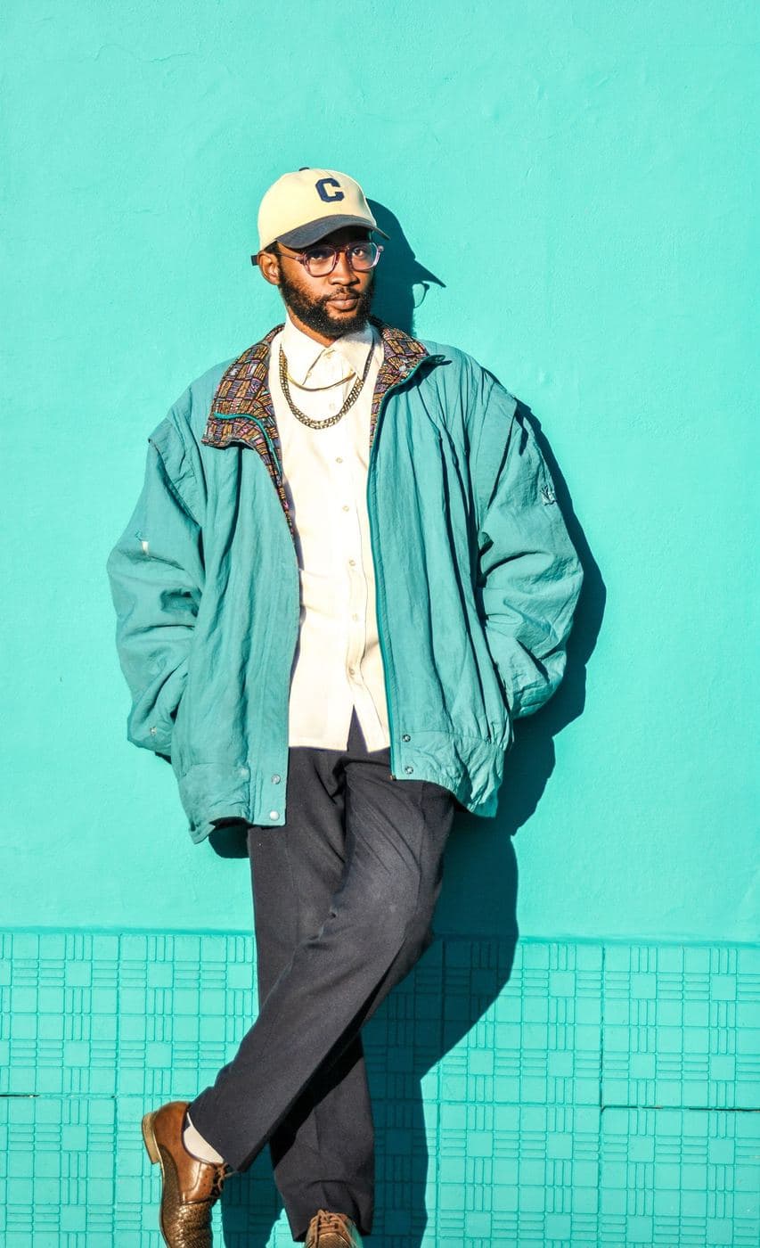 Man in a cap and a teal zip jacket posing against a turquoise tiled wall