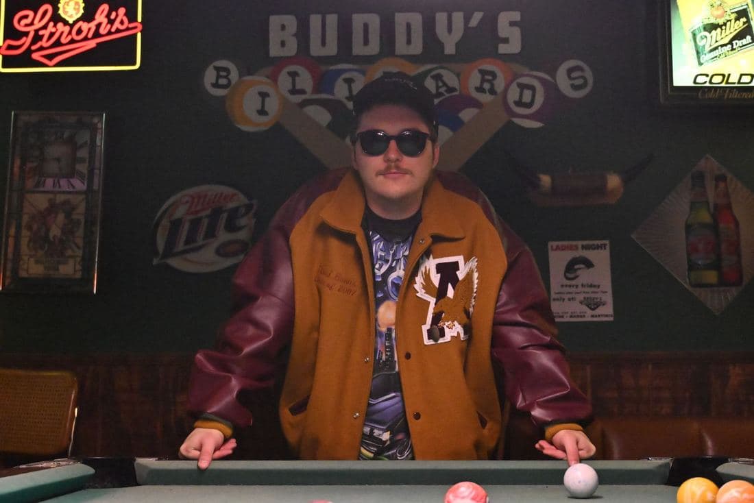 Man in sunglasses and a mustard and burgundy varsity jacket leaning on a pool table